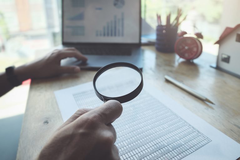 Business man using magnifying to review balance sheet annual with using  laptop computer to calculating budget. audit and Check integrity before investment concept.